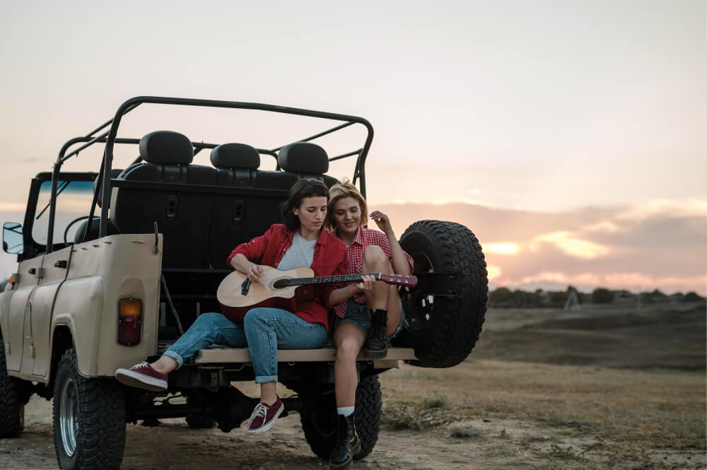Two travelers sitting on a safari jeep at sunset, playing guitar and relaxing during a safari adventure in Tanzania.