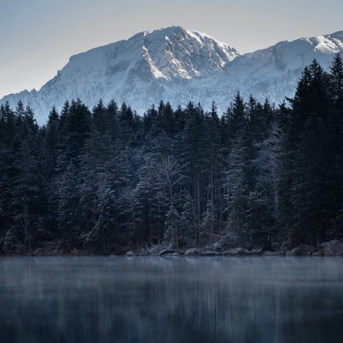 Snow-covered mountain peak reflected on a calm lake with dense pine forest in winter.